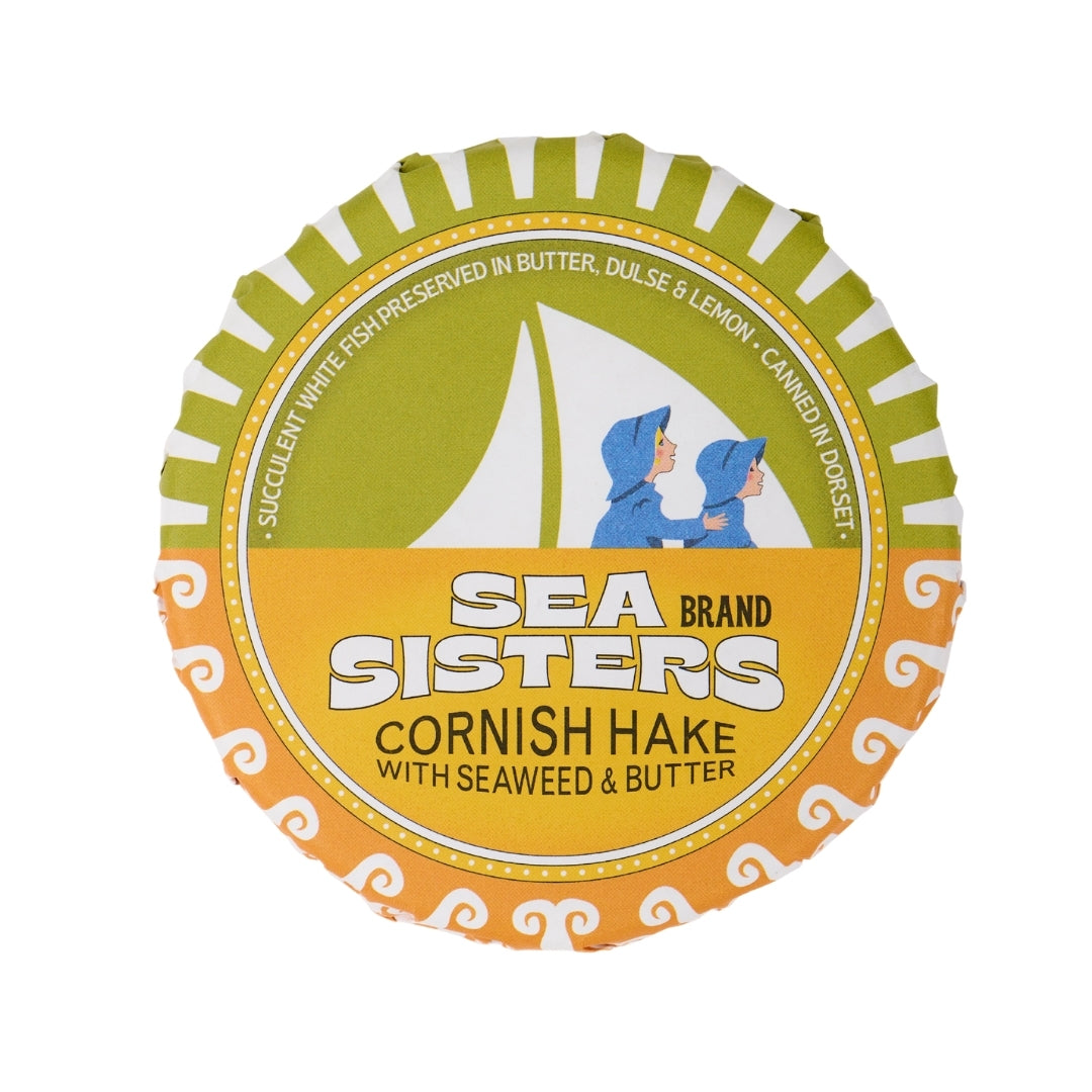 A round Sea Sisters Cornish Hake in Butter package, featuring a green, yellow, and orange label with a white sailboat illustration and two children in blue hoodies.